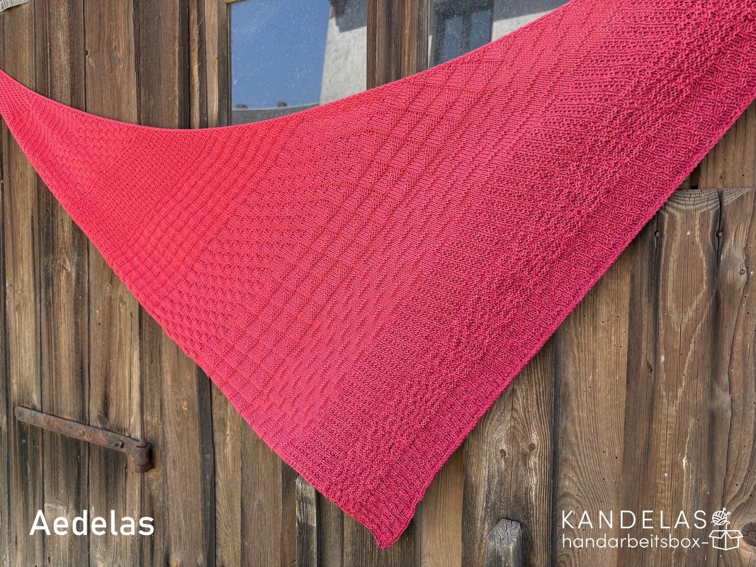 Leuchtend pinkes gestricktes Dreieckstuch mit diagonalem Strukturmuster vor einer Holztür.