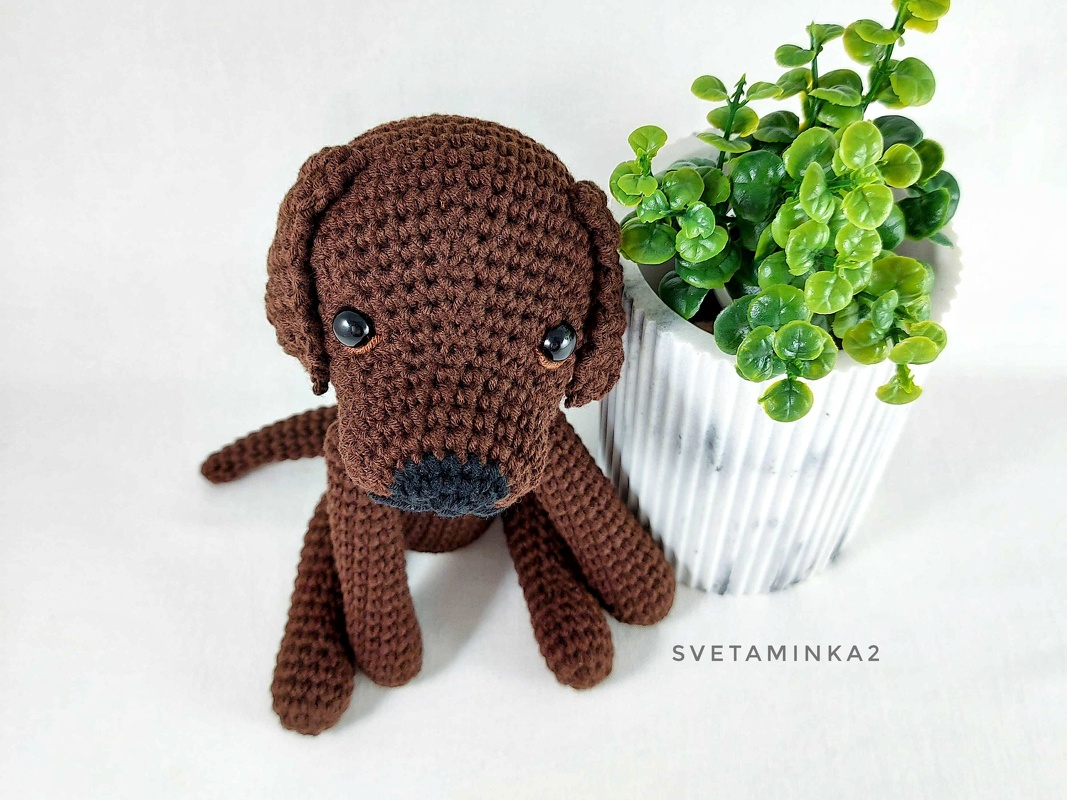 Brown amigurumi dog with long legs and rounded snout on a white background next to a small plant