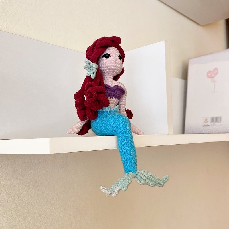 Hand-crocheted mermaid amigurumi doll with red curly hair, purple shell top and blue tail sitting on a white shelf