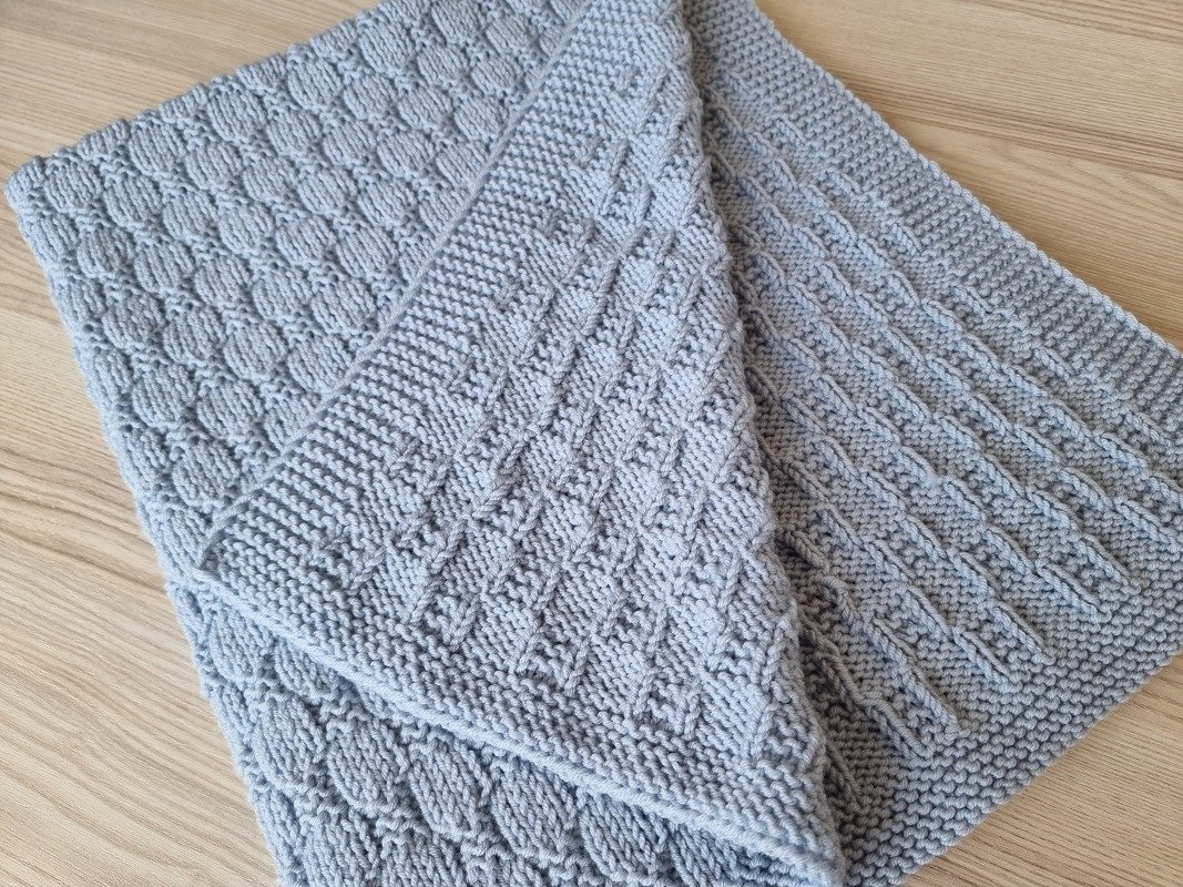 Gefaltete hellgraue gestrickte Babydecke mit Strukturfeldern und Zopfmuster auf hellem Holzhintergrund