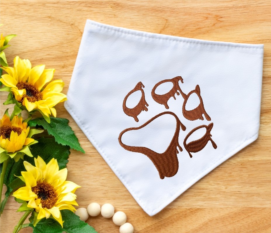 Weißes Dreieckshalstuch mit braun gestickter Pfote mit tropfender Optik auf Holzuntergrund neben Sonnenblumen