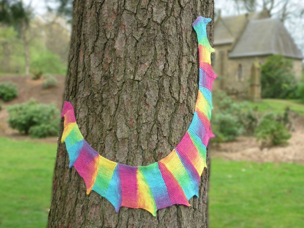 Knitted rainbow dragon tail scarf wrapped around a tree trunk, with a blurred building and lawn in the background.