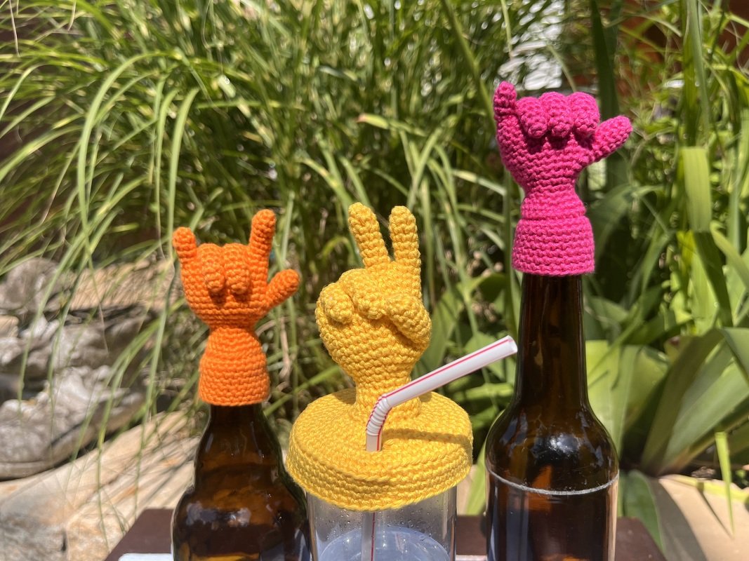 Drei gehäkelte Handgesten‑Aufsätze: orange Rock‑Geste, gelbe Peace‑Geste auf Becher mit Strohhalm, pinke Shaka‑Geste.