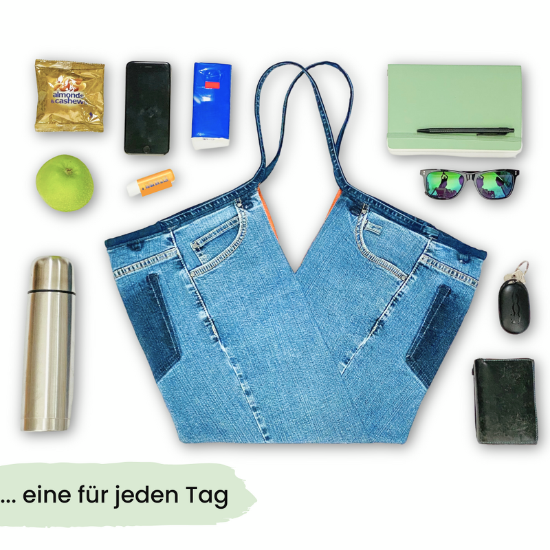 Jeans‑Einkaufstasche im Upcycling‑Look mit V‑Schnitt, flach gelegt und umgeben von typischen Alltagsgegenständen wie Thermosflasche, Apfel, Sonnenbrille, Geldbörse und Handy
