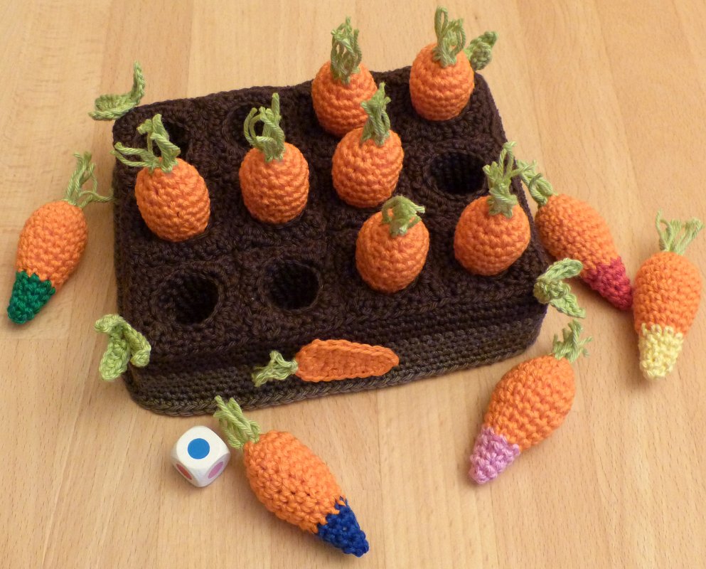 Braune Steckbox mit Löchern, mehrere gehäkelte orange Karotten mit grünen Blättern und ein Würfel mit farbigen Punkten auf hellem Tisch