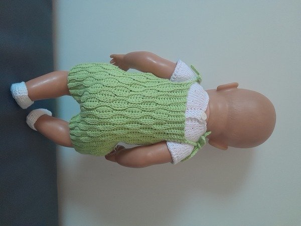 Babypuppe von hinten in grünem gehäkelten Einteiler mit Wellenmuster, weißen Ärmeln und weißen Schuhen