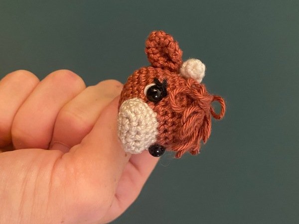 Miniature crocheted reddish-brown pony head amigurumi with light snout and black eyes, held between fingers against a plain background.