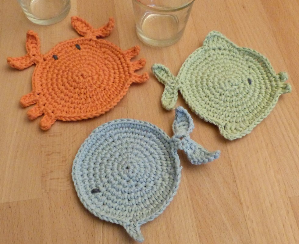 Drei gehäkelte Untersetzer mit Meerestier-Motiven (Krebs, Fisch, Wal) in Pastellfarben auf Holzoberfläche