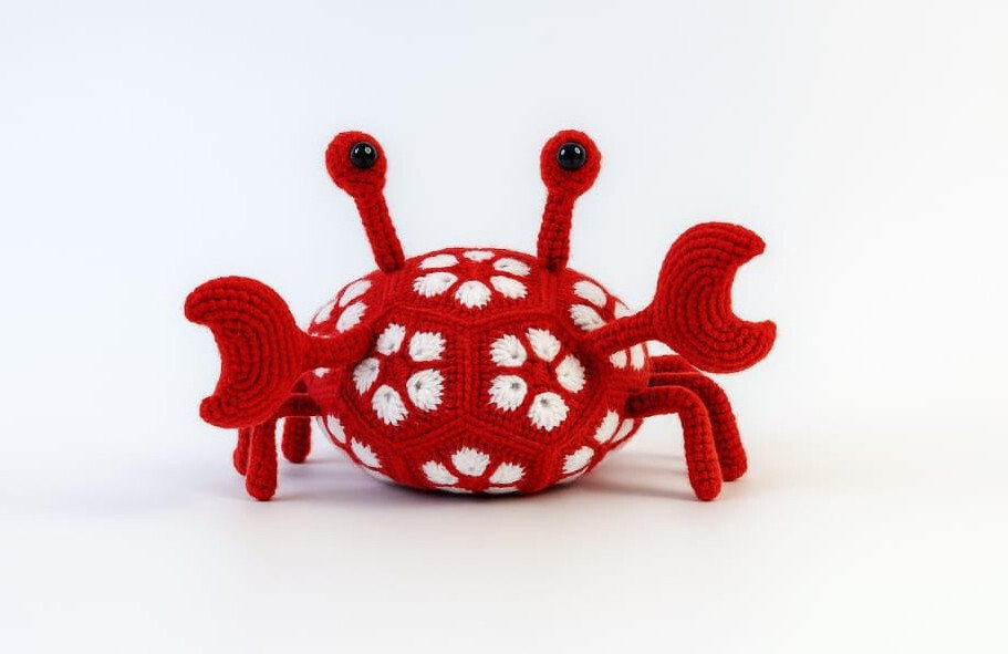 Rotes, gehäkeltes Amigurumi in Krabbenform mit weißen African-Flower-Motiven und schwarzen Augen auf weißem Hintergrund.