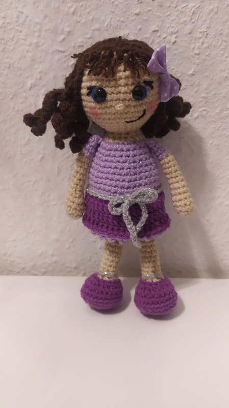 Gehäkelte Amigurumi-Puppe mit lila Kleid, braunen Locken und Schleife