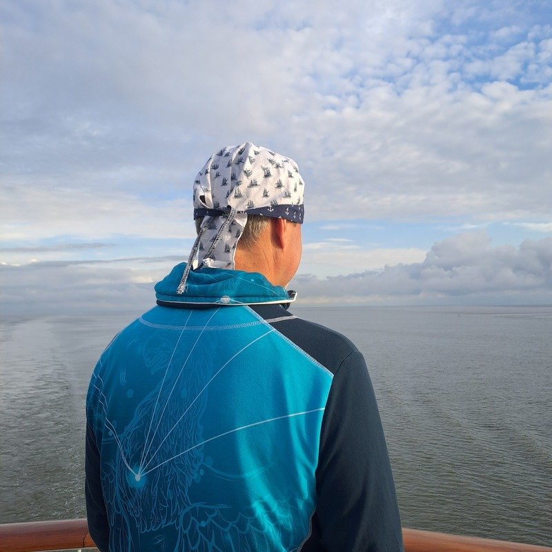 Person von hinten mit gebundenem Bandana mit Segelboot‑Muster vor offenem Meer