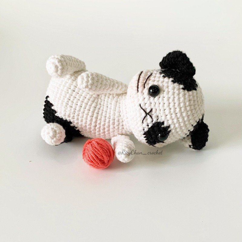 Black-and-white crocheted amigurumi puppy with safety eyes and stitched 'X' mouth beside a coral yarn ball.