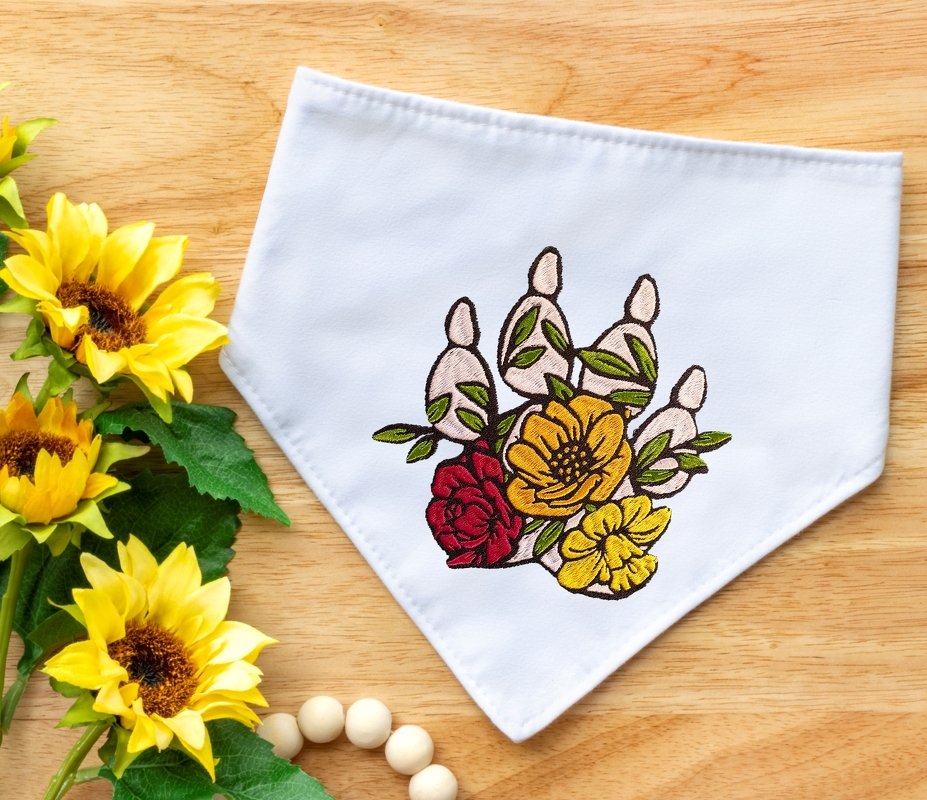 Bestickte Tierpfote mit roten und gelben Blumen auf weißem Stoff, daneben Sonnenblumen und Holzperlen.