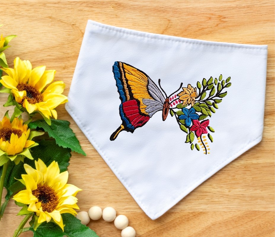 Bunte Stickerei eines Schmetterlings mit Blumen auf weißem Stoff, mit Sonnenblumen und Holzperlen daneben.