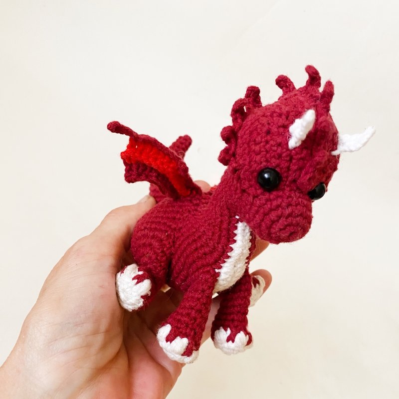 Hand holding a small red crocheted amigurumi dragon with white horns, belly, and claws.