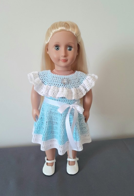 Puppe trägt ein hellblau-weißes gehäkeltes Kleid mit Rüschen, weißer Schleife und weißen Schuhen