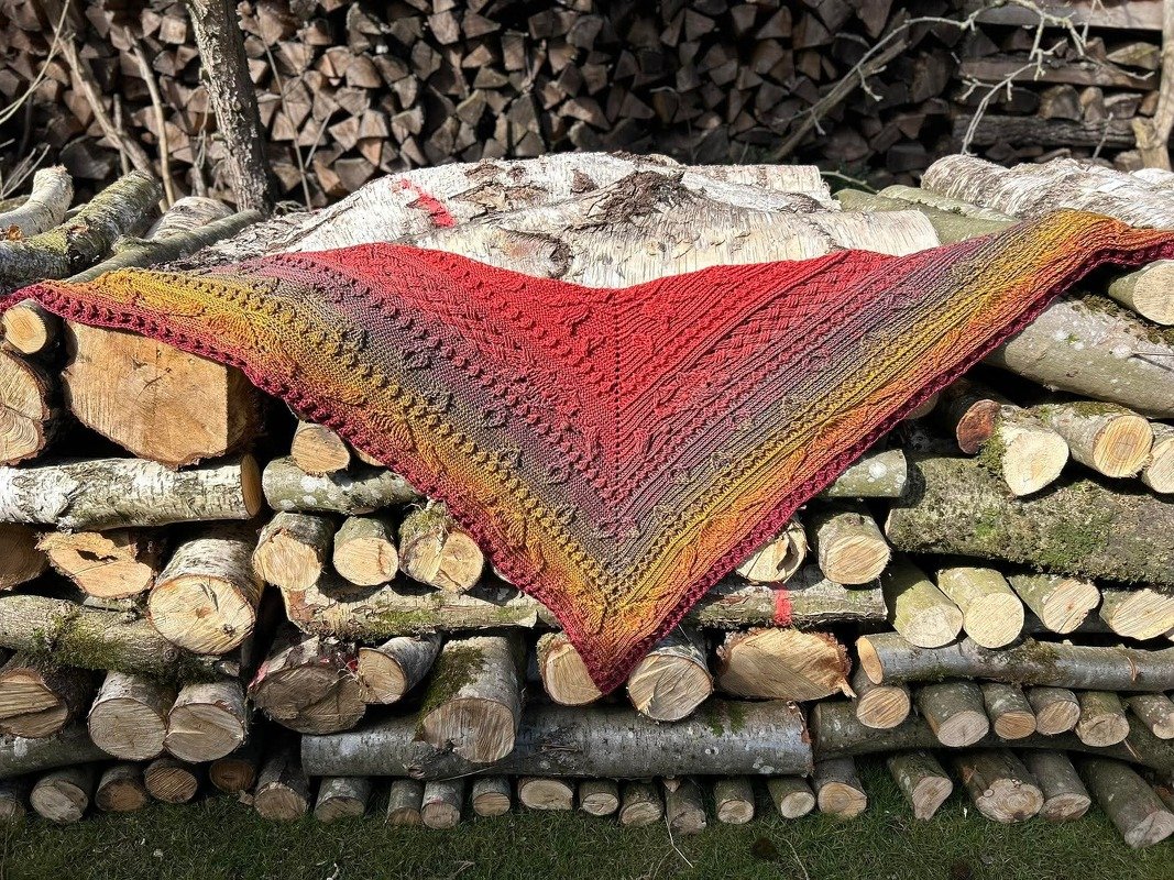 Gehäkeltes Dreieckstuch in Rot-, Orange- und Gelbtönen mit Strukturmuster auf einem Holzstapel.