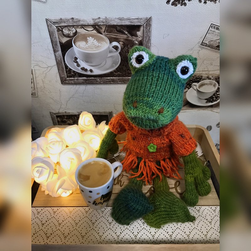 Gestrickter grüner Amigurumi-Frosch mit weißen Augen und orangefarbenem Kleid neben Kaffeetasse und Lichterrosen
