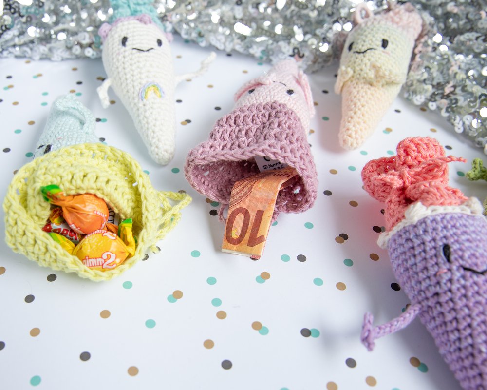 Mehrere schnell gehäkelte Amigurumi-Täschchen in Pastellfarben, einige geöffnet und mit Süßigkeiten oder Geldscheinen gefüllt, auf festlichem Konfetti-Hintergrund.