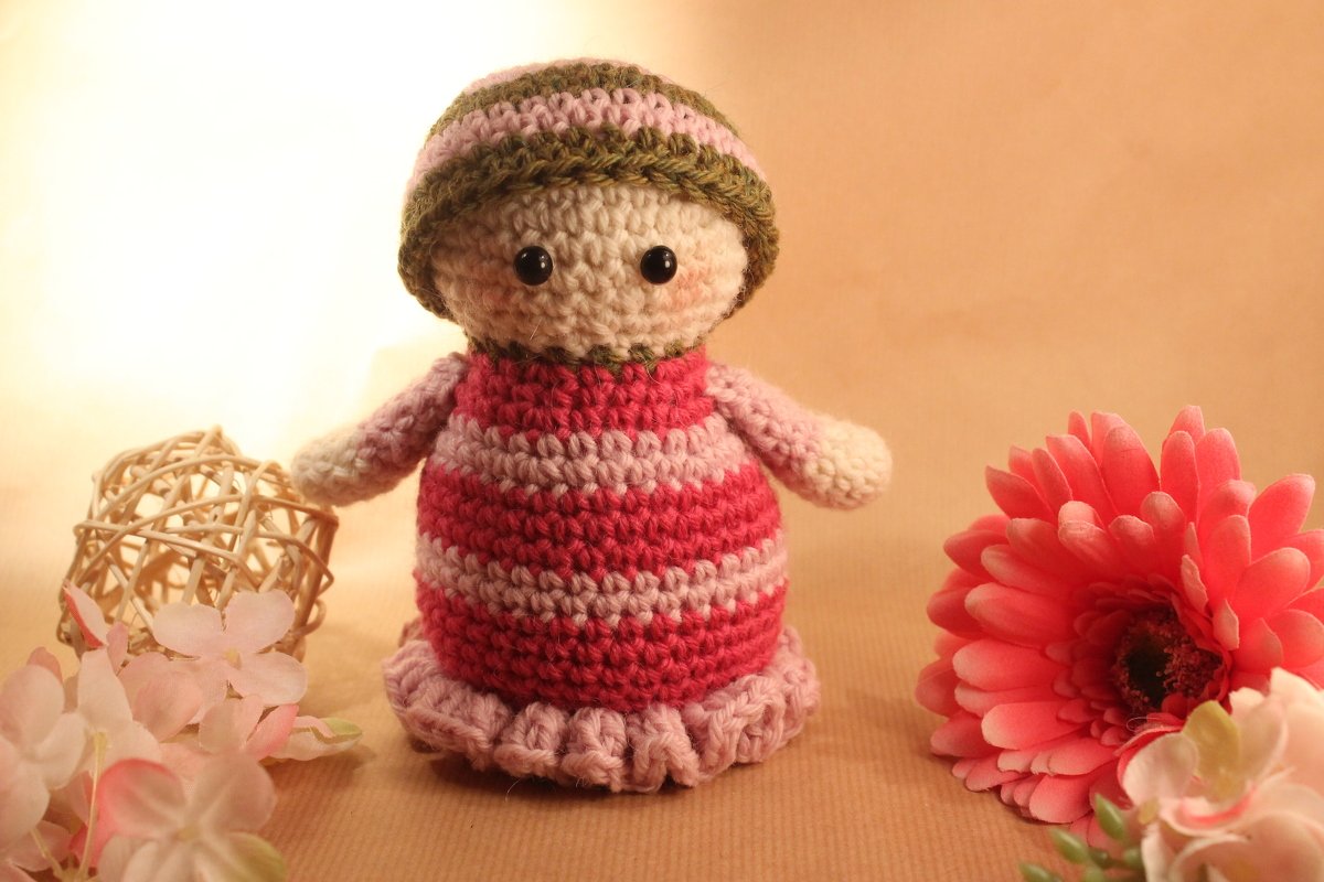 Gehäkelte Amigurumi‑Puppe mit gestreiftem Kleid und Mütze, umgeben von Dekoblumen