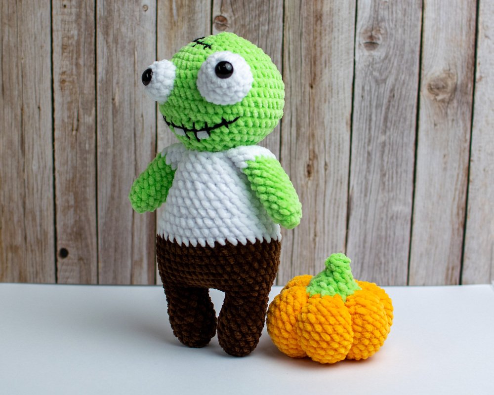 Gehäkelte grüne Amigurumi-Puppe mit kleinem orangefarbenem Kürbis vor Holzwand