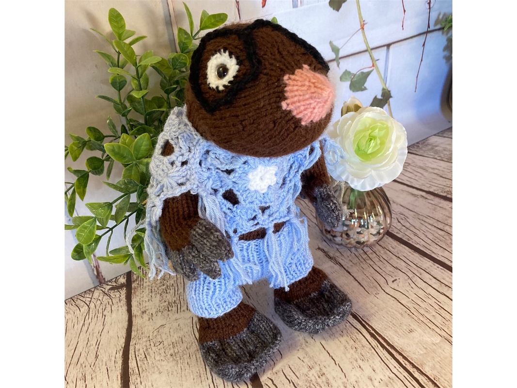 Handgestrickter brauner Maulwurf mit rosafarbener Nase und hellblauem Outfit auf Holzdielen vor Pflanzen.