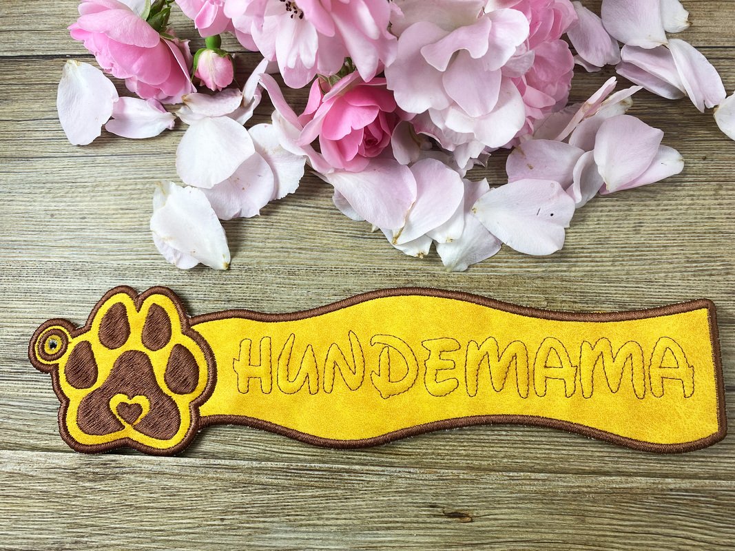 Gelb-brauner bestickter Schlüsselanhänger 'HUNDEMAMA' mit Pfoten-Herz auf Holz, umgeben von rosa Rosenblättern.