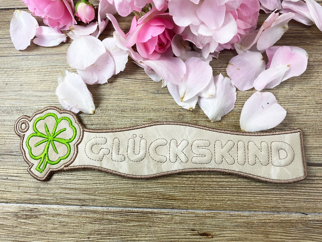 Länglicher beiger ITH‑Schlüsselanhänger mit gesticktem Schriftzug GLÜCKSKIND und grünem Kleeblatt auf Holz
