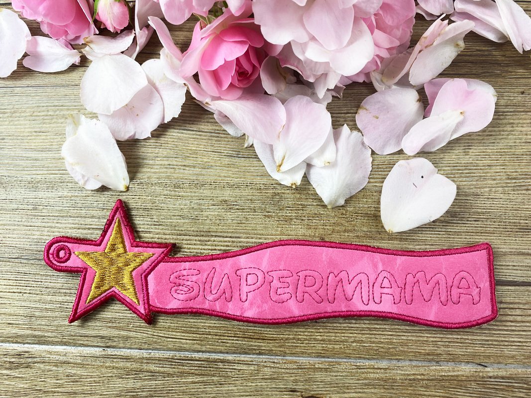 Rosa ITH-Schlüsselanhänger 'SUPERMAMA' mit goldfarbenem Stern auf Holzhintergrund und Rosenblüten.