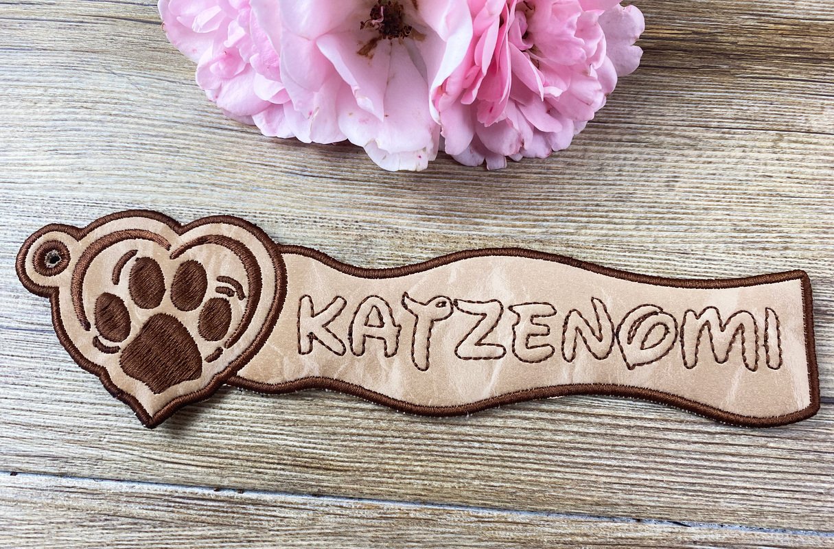 Bestickter Schlüsselanhänger aus hellbeigem Material mit Pfotenherz und dem Schriftzug 'KATZENOMI' auf Holzuntergrund, daneben rosa Blüten.