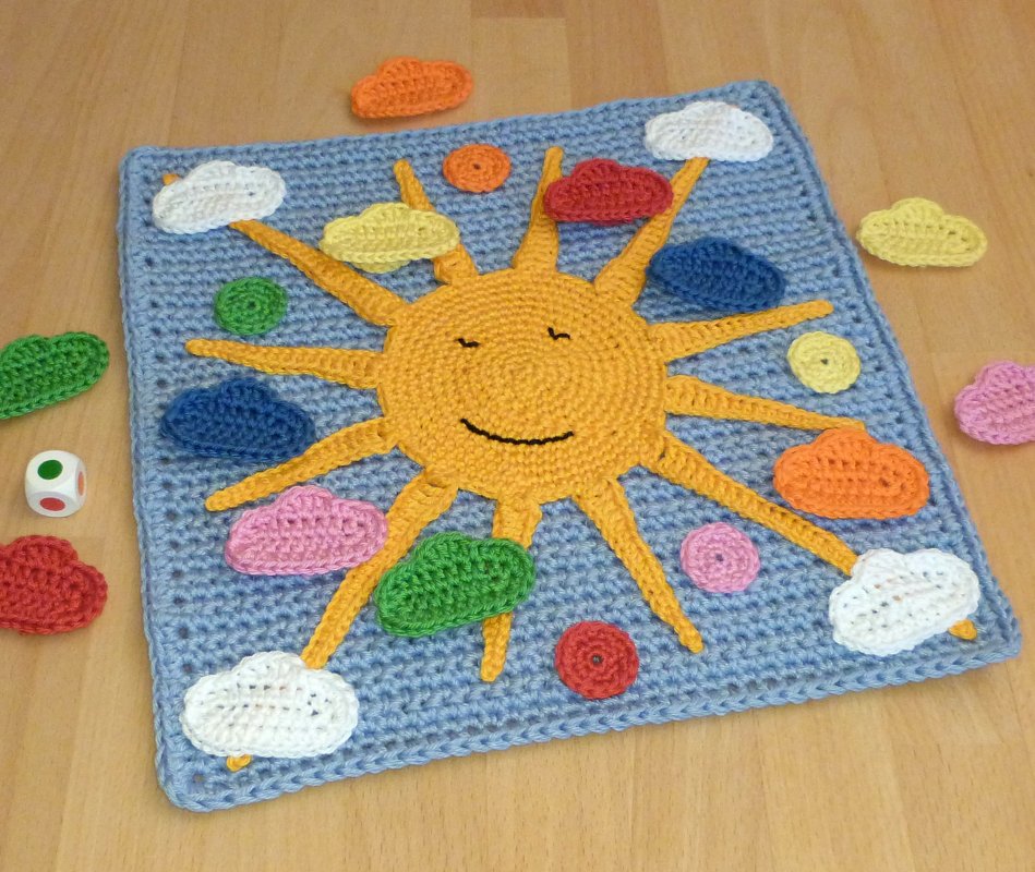 Gehäkeltes Spielbrett mit gelber Sonne, bunten Wolken und einem Würfel auf Holzboden.