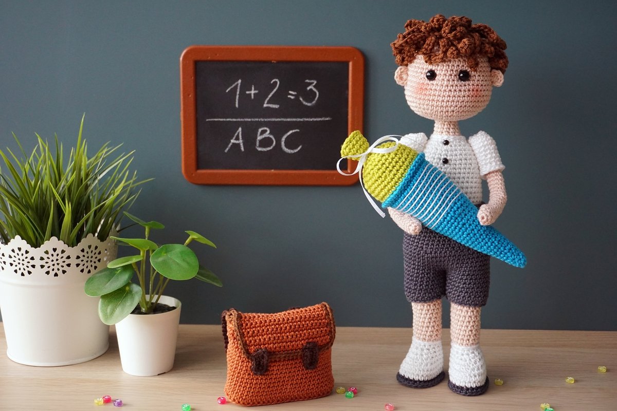 Gehäkelter Amigurumi-Junge in weißem Hemd und dunkler Hose, der eine blau-gelbe Schultüte hält; im Hintergrund eine Tafel mit '1+2=3' und 'ABC' sowie ein gehäkelter Ranzen.