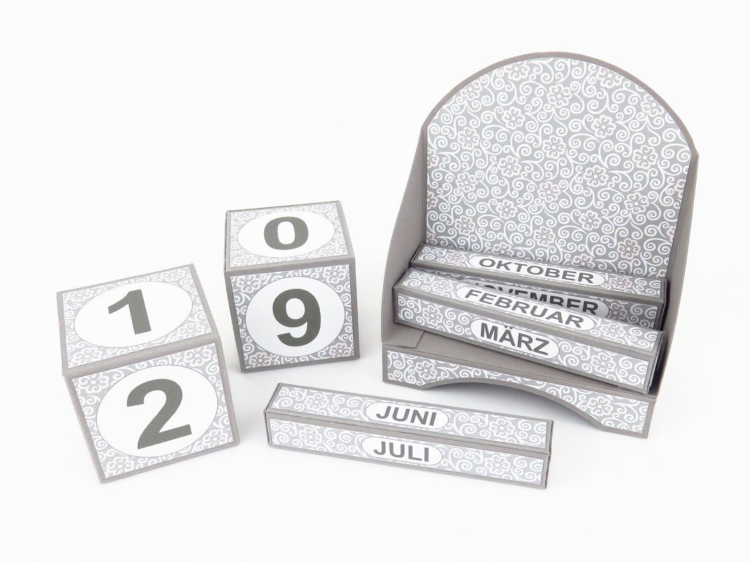 Bastelprojekt für einen ewigen Papierkalender: zwei Zahlenwürfel mit Ziffern und beschriftete Monatsstreifen in grau-weißem Ornamentmuster
