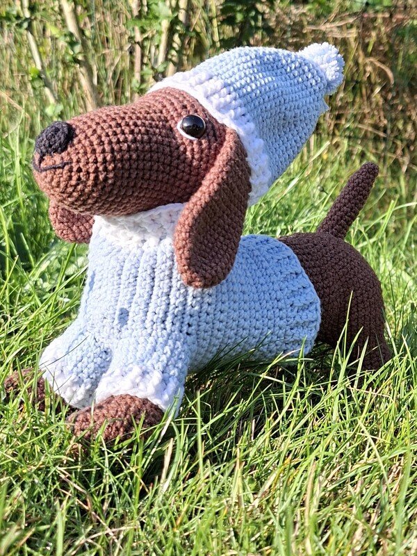 Gehäkelter Dackel-Amigurumi mit hellblauem Pullover und Mütze auf Gras