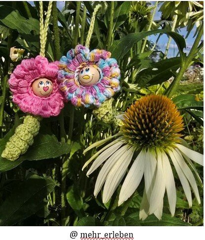 Nahaufnahme zweier gehäkelter Blumen-Anhänger mit bemalten Gesichtern, hängend an grünen Stielen