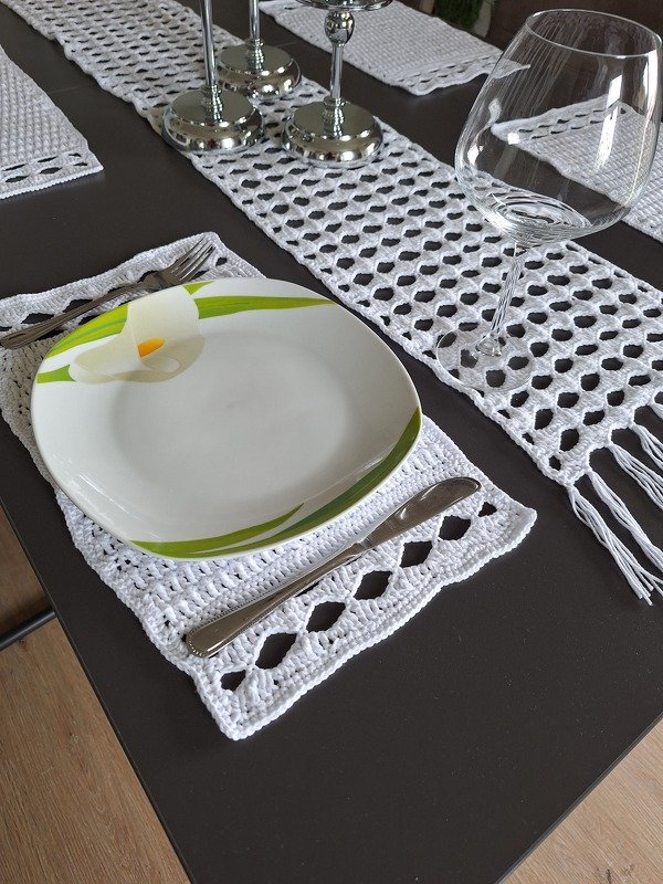 Weißer gehäkelter Tischläufer und Platzdeckchen mit Netzmuster auf dunklem Tisch, Teller, Besteck und Weinglas