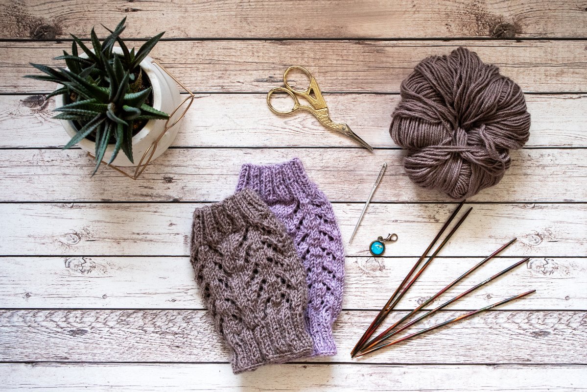 Zwei gestrickte Kinder-Beinstulpen, Wollknäuel, Stricknadeln und Schere auf hellem Holztisch.