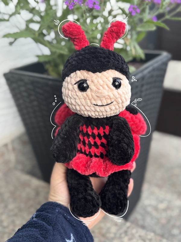 Hand hält gehäkeltes Marienkäfer-Amigurumi in Schwarz und Rot mit beiger Gesichtsfläche und schwarzen Augen, Pflanzenkübel im Hintergrund