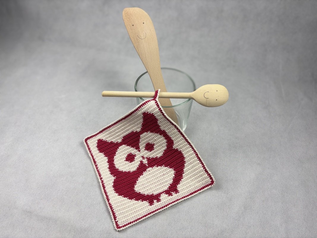 Quadratischer zweifarbiger Topflappen mit Eulen-Grafik und schmaler Borte, auf hellgrauem Hintergrund neben Küchenlöffeln