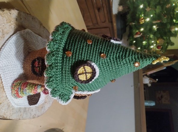 Gehäkelter grüner Weihnachtsbaum mit Perlen und rundem Fensterakzent neben einem gehäkelten Lebkuchenhaus auf einer Holzscheibe; im Hintergrund ein beleuchteter Weihnachtsbaum.