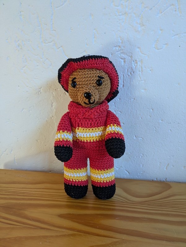 Gehäkelter Amigurumi-Teddybär in roter Feuerwehruniform mit gelb-weißen Streifen und Helm