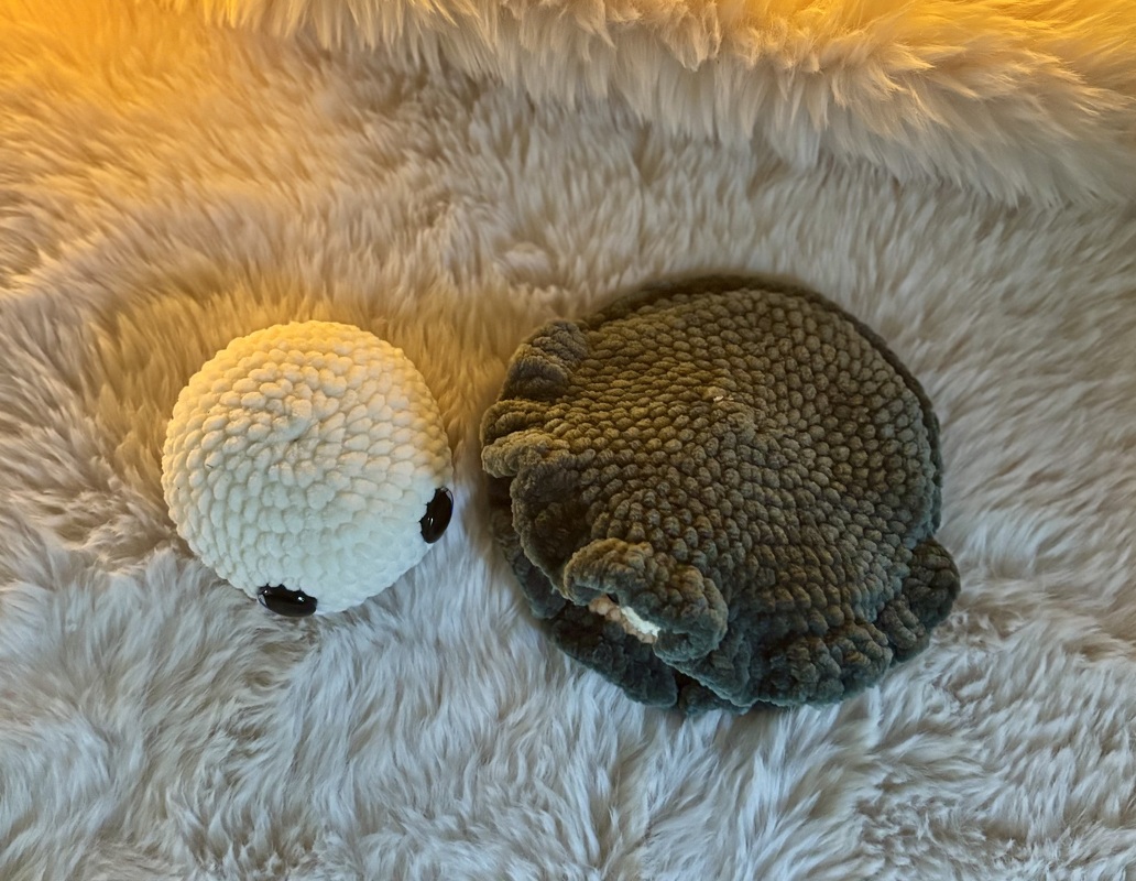 Zwei gehäkelte Amigurumi: weißes Kugelwesen mit Augen und graugrünes, gerüschtes Meereswesen auf flauschigem Untergrund