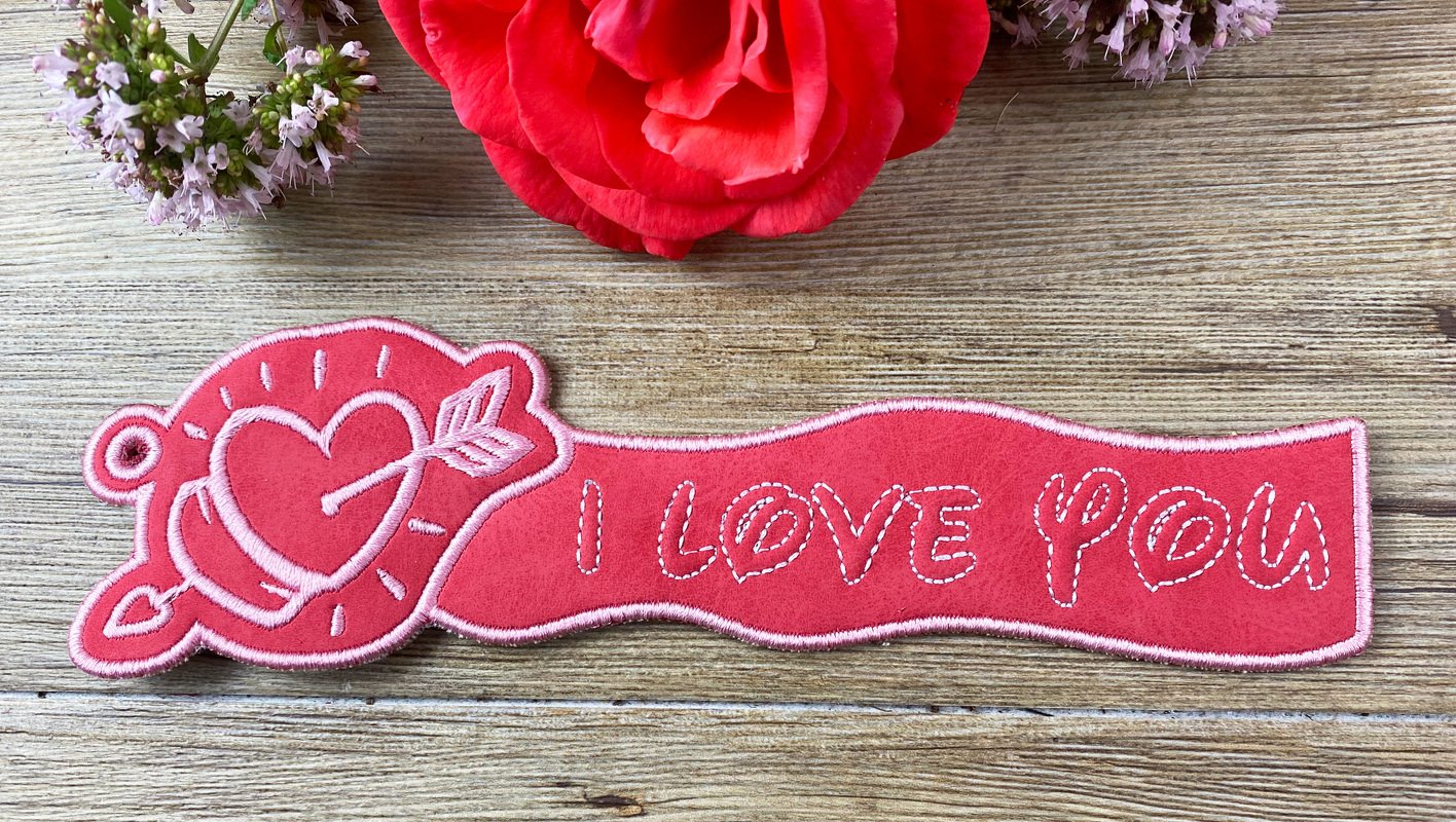 Roter ITH‑Schlüsselanhänger mit Herz, Pfeil und gesticktem Schriftzug ‚I LOVE YOU‘ auf Holzuntergrund
