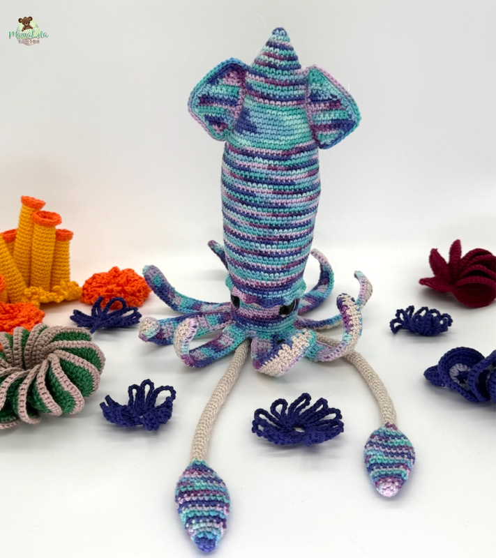 Gehäkelter Amigurumi-Tintenfisch in Blau-, Lila- und Grüntönen, umgeben von gehäkelten Korallen