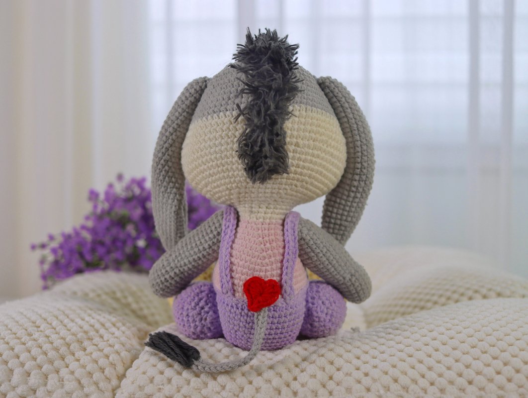 Gehäkelter Esel (Amigurumi) von hinten auf cremefarbener Decke mit lila Latzhose und rotem Herz