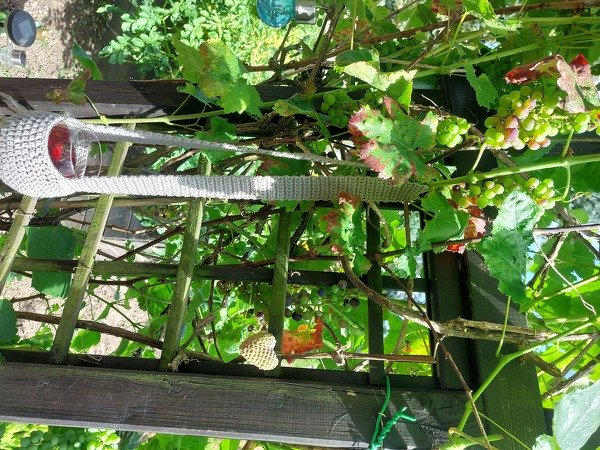 Helle gehäkelte Glashülle an Holzgitter mit Weinreben und kleinem Herzanhänger