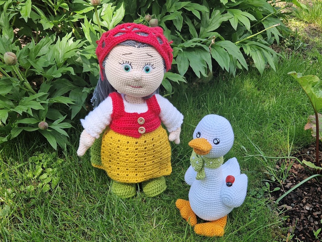 Gehäkelte Amigurumi-Puppe mit rotem Haarnetz, roter Weste und gelbem Rock neben einer weißen Ente mit grünem Schal auf Rasen