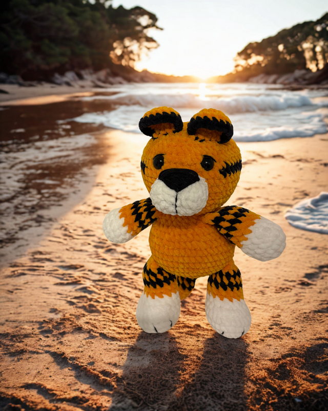 Gehäkelter Amigurumi‑Tiger mit orange‑schwarzen Streifen auf Sand vor Sonnenuntergang