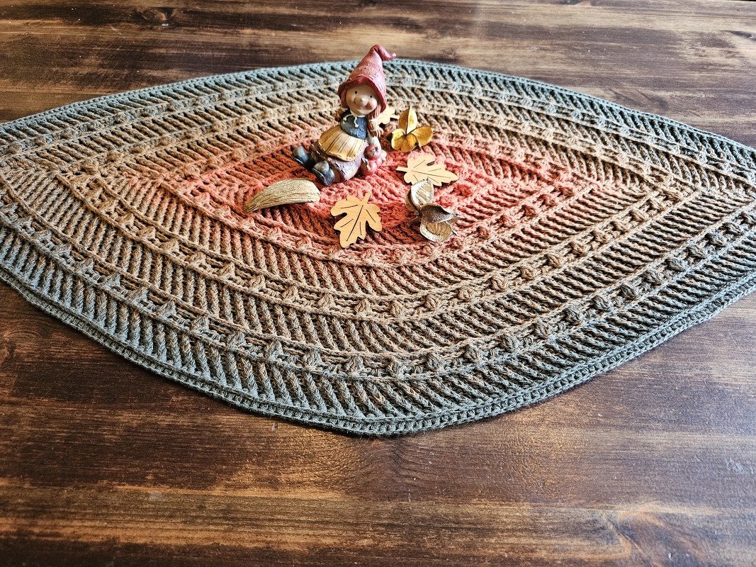 Gehäkelter ovaler Tischläufer mit warmem orangefarbenem Zentrum und grauem Rand, dekoriert mit kleiner Wichtelfigur und Holzblättern auf Holztisch
