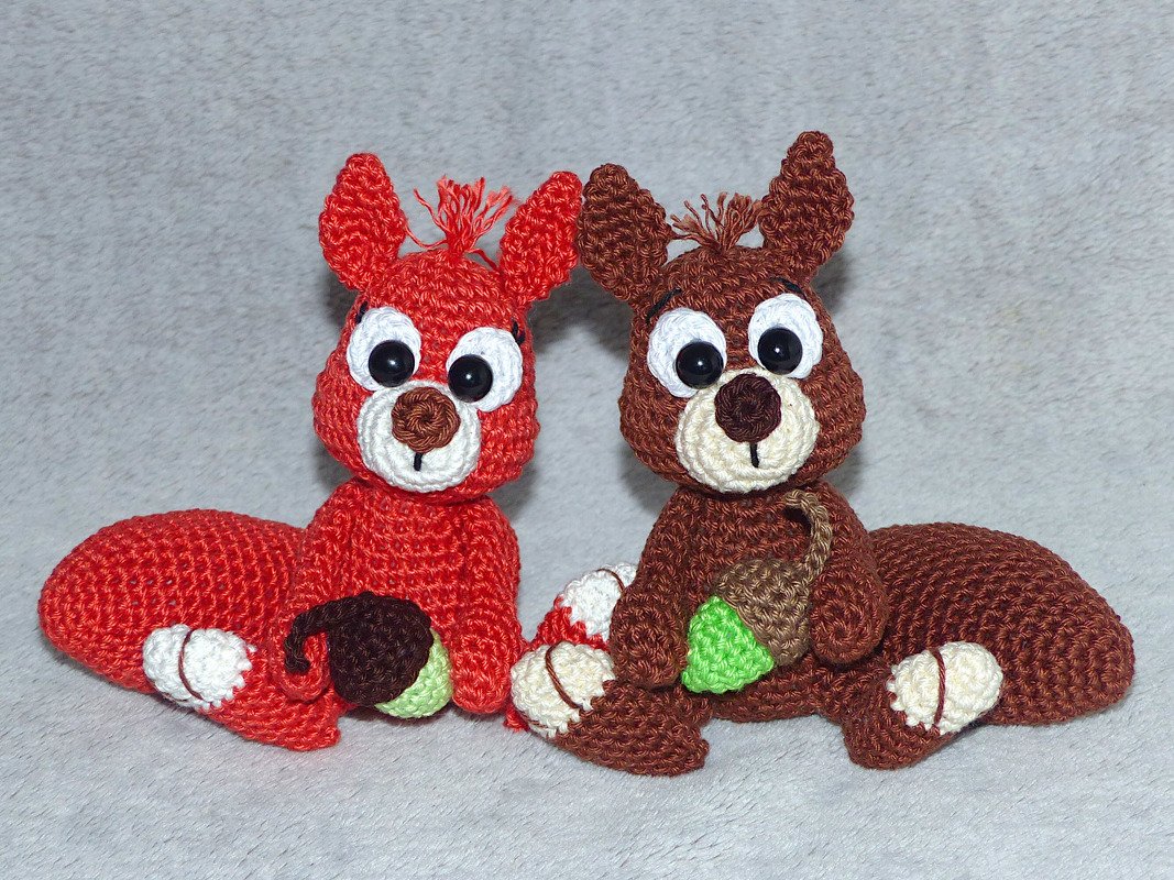 Zwei gehäkelte Amigurumi‑Eichhörnchen, rot und braun, halten jeweils eine Eichel.